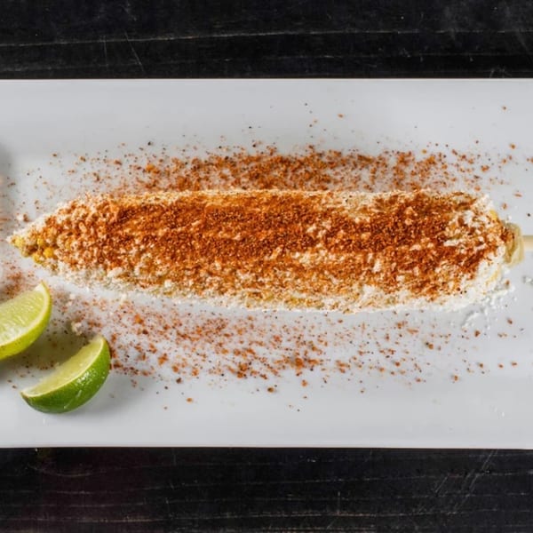 Elote, Mexican street corn, on a white plate with chili powder, cheese, and lime wedges.