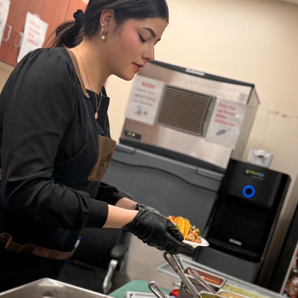 A Casa Nueva employee in a black apron and gloves prepares tacos at a food station.