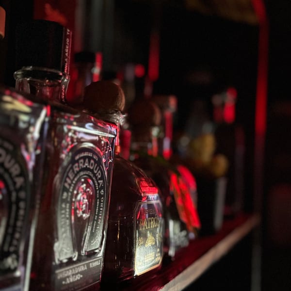 Close-up of tequila bottles, including Herradura and Patron, on a dimly lit bar shelf with red lighting.