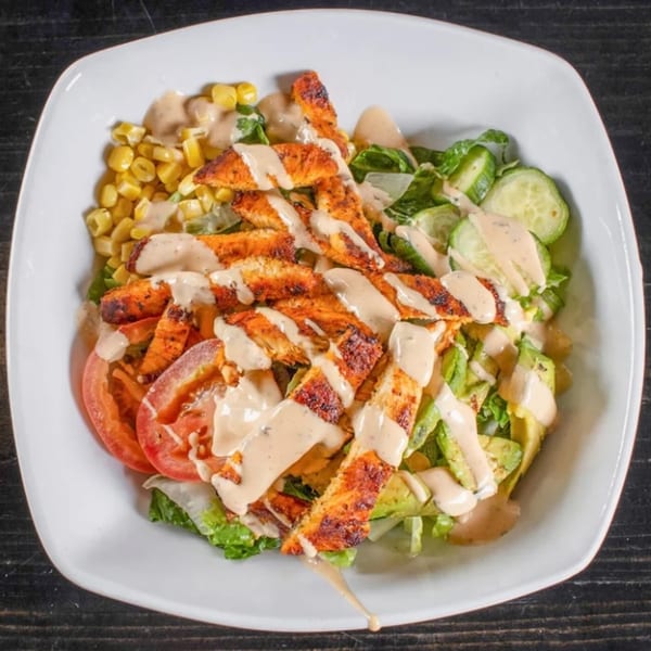 Grilled chicken salad with corn, tomatoes, cucumbers, avocado, and creamy dressing in a white bowl.