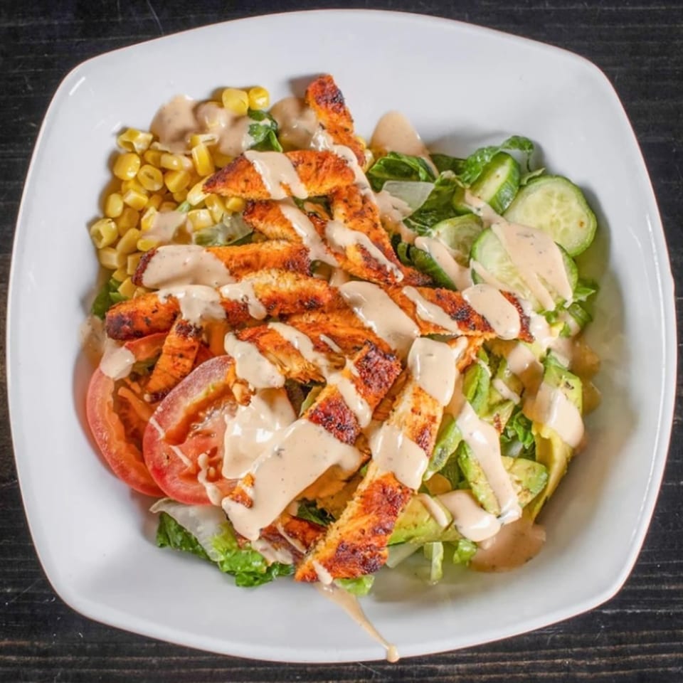 Grilled chicken salad with corn, tomatoes, cucumbers, avocado, and creamy dressing in a white bowl.