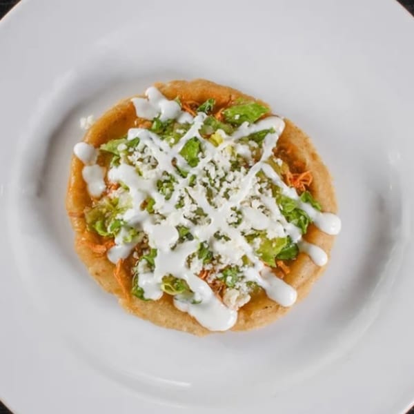 A single Casa Nueva Mexican sopes topped with shredded chicken, lettuce, crumbled cheese, and crema.