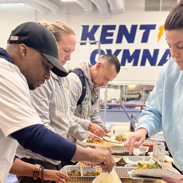 People serving themselves Mexican food from aluminum trays at a buffet line in a gymnasium.